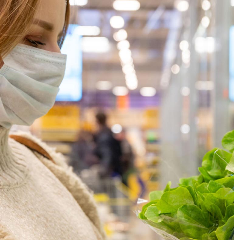 Photography: The image has a blurred background with light bulbs on the roof of a supermarket. In the foreground, the front part of a woman's face with blond hair covering part of her eye. The woman wears a white mask for easy protection because of Covid-19. She wears a white woolen blouse and the handle of a brown leather bag is noticeable over her left shoulder. She observes lettuce leaf, which appear in the lower right corner of the image