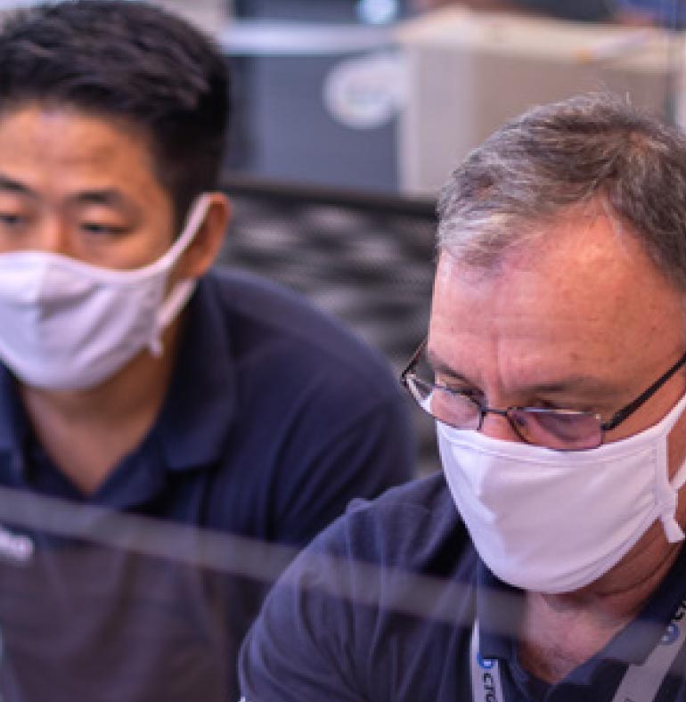 Photography: Two men are sitting next to each other, looking at an object that does not appear in the photograph. The one on the left, further down, has oriental physical characteristics and black hair and uses a white mask to cover the nose and mouth. The one on the right is a Caucasian man with gray hair and bald head. He wears glasses and a white mask to cover his nose and mouth. A gray badge strip with the printed blue CTG logo is seen