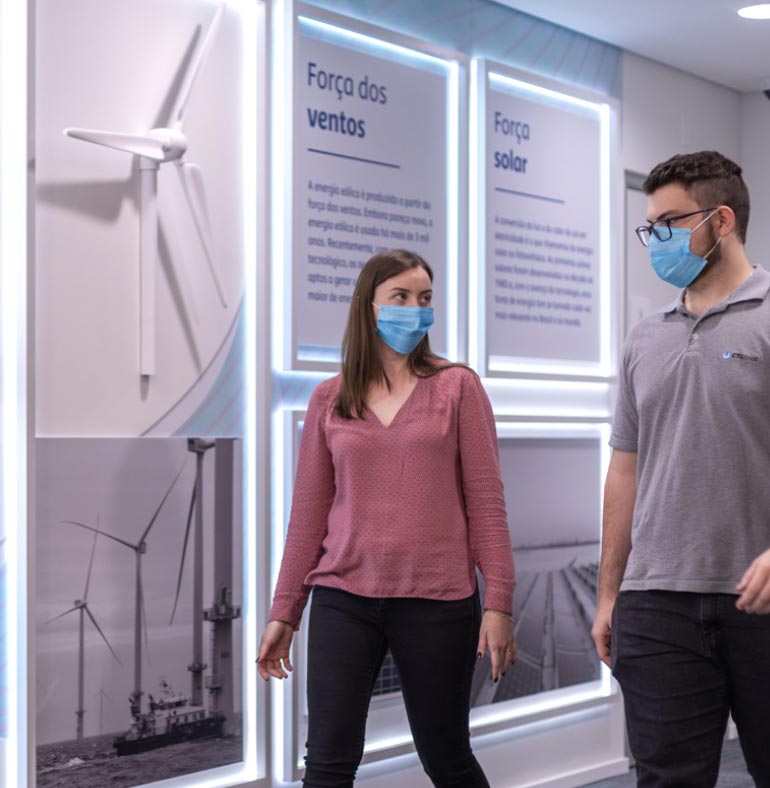 Photography: The photograph shows a wall decorated with black and white pictures of wind blades and two illuminated panels that read the phrases wind force and solar force. In front of this wall, a woman and a man walk side by side, looking at each other, apparently talking. Both use a blue mask to cover their nose and mouth. The woman wears a pink blouse and black pants and the man, wearing glasses, wears a gray polo shirt and black pants