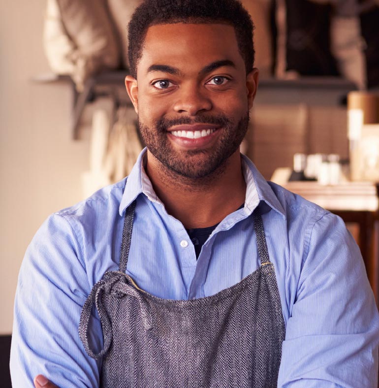 Photography: Black man smiles looking at the camera. He has dark hair and a beard on his face. He is dressed in a light blue shirt and has a gray work apron