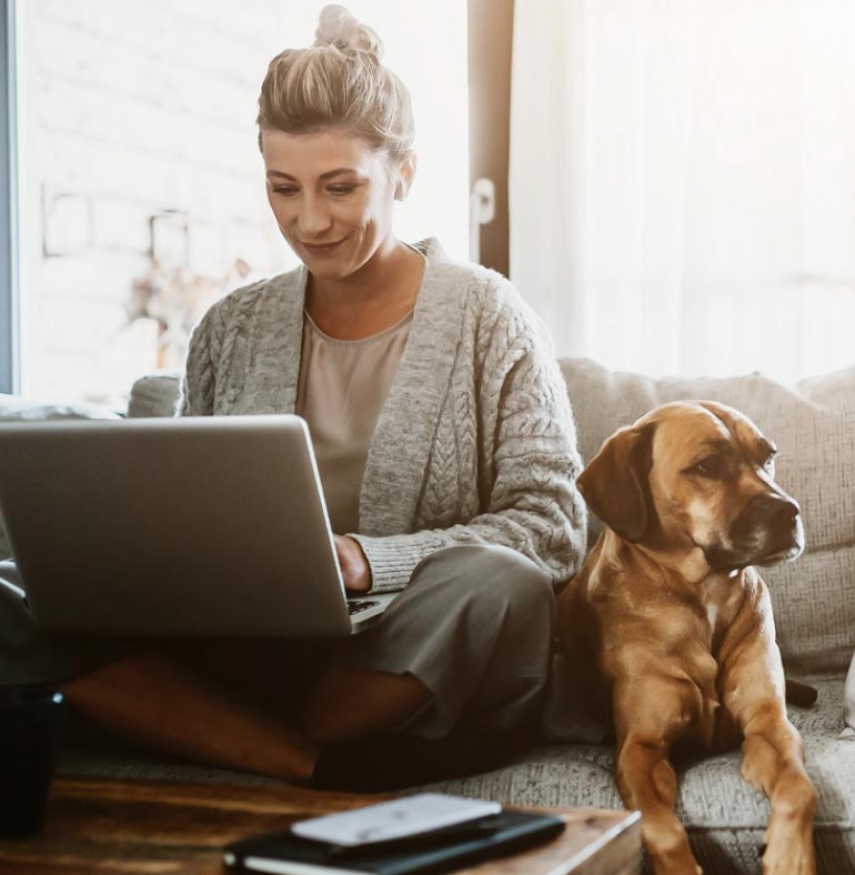 Fotografia: mulher loira está sentada com as pernas cruzadas sobre o sofá cinza da sala e notebook prateado sobre o colo. Ela olha para o computador sorridente. Ao seu lado, sobre o sofá, um cachorro marrom de pelos curtos e nariz preto está encostado em sua perna. No canto inferior, aparece a ponta de uma mesa de madeira com agenda preta e celular apoiados sobre ela