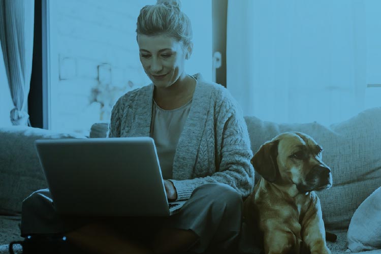 Fotografia: mulher loira está sentada com as pernas cruzadas sobre o sofá cinza da sala e notebook prateado sobre o colo. Ela olha para o computador sorridente. Ao seu lado, sobre o sofá, um cachorro marrom de pelos curtos e nariz preto está encostado em sua perna. No canto inferior, aparece a ponta de uma mesa de madeira com agenda preta e celular apoiados sobre ela