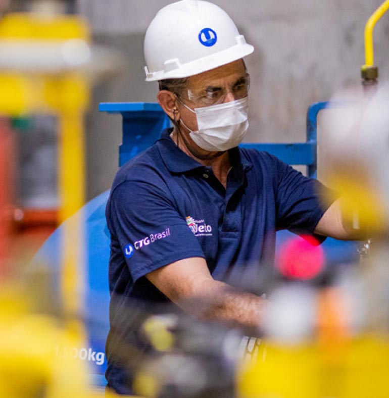 Fotografia: funcionário da CTG Brasil observa e trabalha sobre equipamento. Ele está usando um capacete de operário branco com o logotipo da empresa na cor azul na frente. Usa óculos de proteção transparente e máscara de proteção facial. Na camisa azul, o logotipo da empresa está estampado na cor branca na manga direita. A fotografia possui tons de amarelo nas laterais, efeito de equipamento que ficou desfocado por estar em primeiro plano