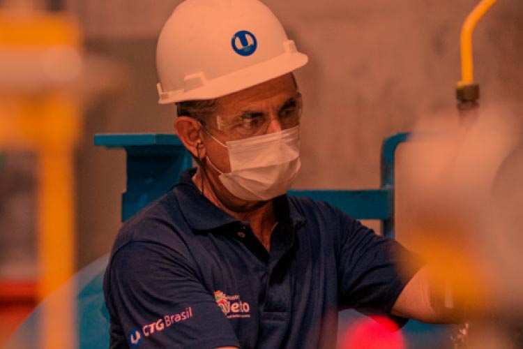 Fotografia: funcionário da CTG Brasil observa e trabalha sobre equipamento. Ele está usando um capacete de operário branco com o logotipo da empresa na cor azul na frente. Usa óculos de proteção transparente e máscara de proteção facial. Na camisa azul, o logotipo da empresa está estampado na cor branca na manga direita. A fotografia possui tons de amarelo nas laterais, efeito de equipamento que ficou desfocado por estar em primeiro plano