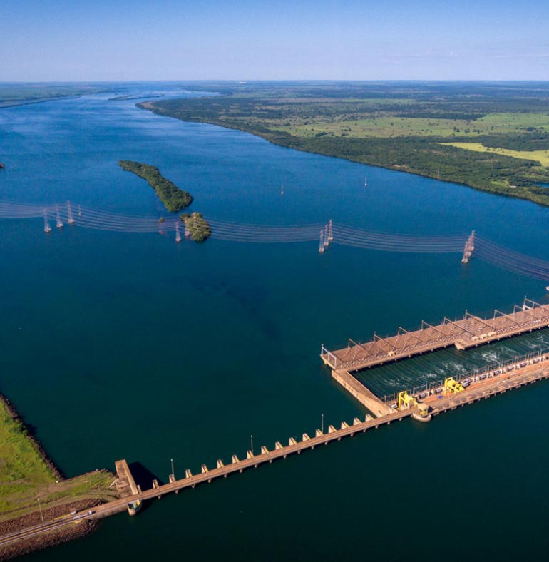 Fotografia Imagem aérea mostra um rio de cor azul largo ladeado por áreas verdes e uma ilha coberta de vegetação no meio. Por cima dessa ilha, passam linhas de transmissão de energia sustentadas por torres construídas no leito do rio. Na parte inferior da foto, vê-se uma barragem de concreto represando o rio. A barragem forma uma área retangular na área inferior direita da foto, onde a água do rio fica mais movimentada