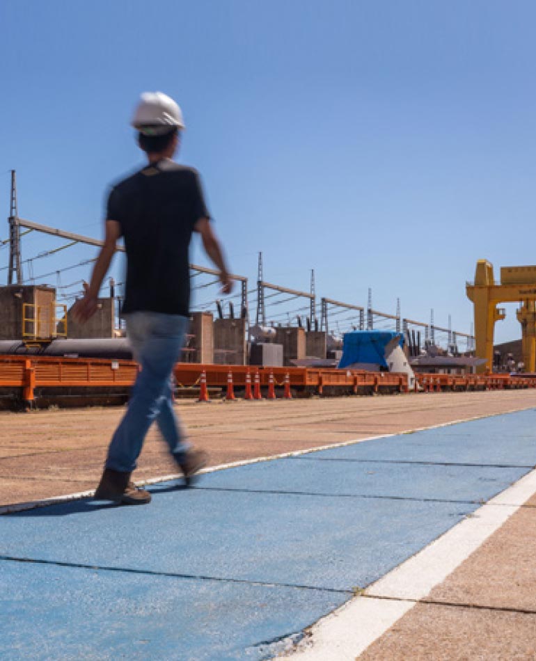 Fotografia: céu azul no fundo da foto. Homem caminha sobre chão de cimento pintado de azul. Do seu lado esquerdo há cones laranjas apoiados no chão e equipamentos de grande porte de usina hidrelétrica. No fundo há um guindaste amarelo