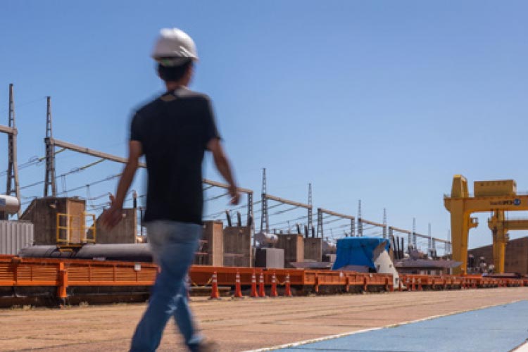 Fotografia: céu azul no fundo da foto. Homem caminha sobre chão de cimento pintado de azul. Do seu lado esquerdo há cones laranjas apoiados no chão e equipamentos de grande porte de usina hidrelétrica. No fundo há um guindaste amarelo