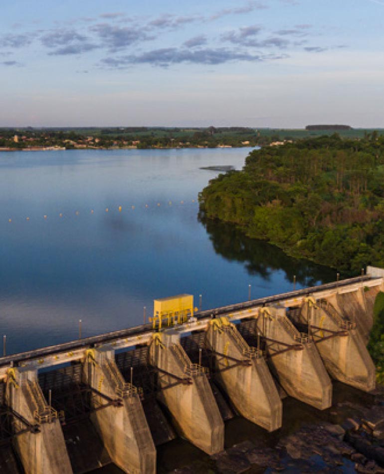 Fotografia: céu azul com algumas nuvens sobre um rio com margem coberta por árvores de grande porte. Na parte inferior da imagem, vê-se a barragem de uma usina