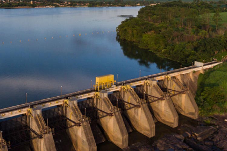 Fotografia: céu azul com algumas nuvens sobre um rio com margem coberta por árvores de grande porte. Na parte inferior da imagem, vê-se a barragem de uma usina