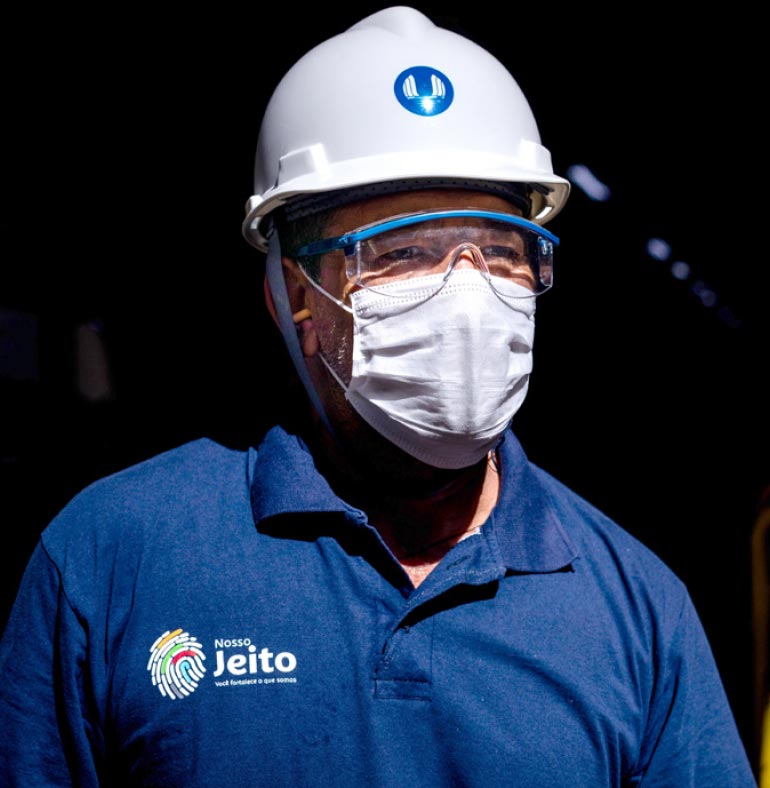 Fotografia: Homem observa o horizonte utilizando um capacete branco com o logotipo da CTG Brasil na cor azul estampado na área frontal. Ele utiliza óculos transparentes de proteção e uma máscara branca que encobre o nariz e a boca. A camisa azul possui um impresso colorido, na altura do peito, do lado direito, que se assemelha a uma impressão digital. Ao lado do desenho está a plavra Jeito, acompanhada de outras palavras ilegíveis na imagem.