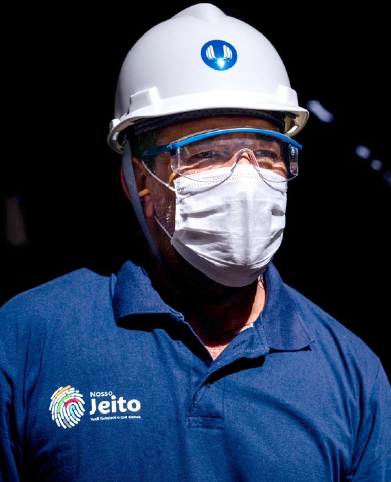 Fotografia: Homem observa o horizonte utilizando um capacete branco com o logotipo da CTG Brasil na cor azul estampado na área frontal. Ele utiliza óculos transparentes de proteção e uma máscara branca que encobre o nariz e a boca. A camisa azul possui um impresso colorido, na altura do peito, do lado direito, que se assemelha a uma impressão digital. Ao lado do desenho está a plavra Jeito, acompanhada de outras palavras ilegíveis na imagem