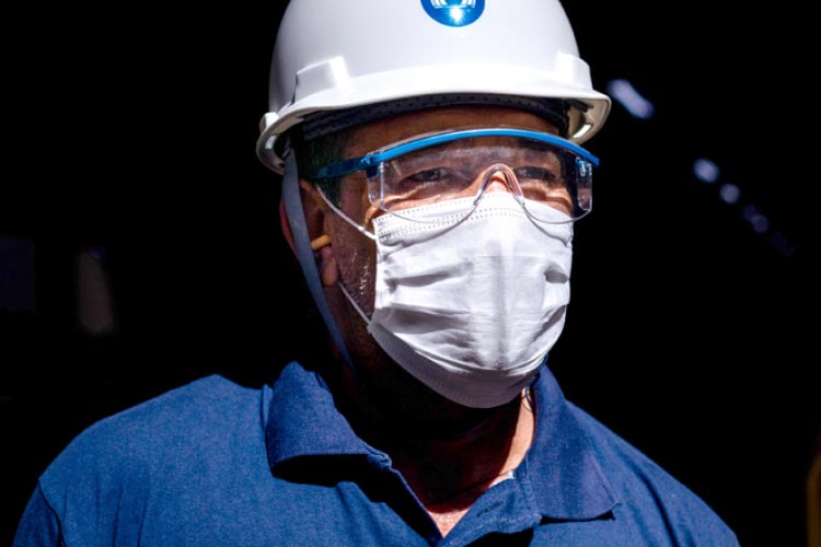 Fotografia: Homem observa o horizonte utilizando um capacete branco com o logotipo da CTG Brasil na cor azul estampado na área frontal. Ele utiliza óculos transparentes de proteção e uma máscara branca que encobre o nariz e a boca. A camisa azul possui um impresso colorido, na altura do peito, do lado direito, que se assemelha a uma impressão digital. Ao lado do desenho está a plavra Jeito, acompanhada de outras palavras ilegíveis na imagem