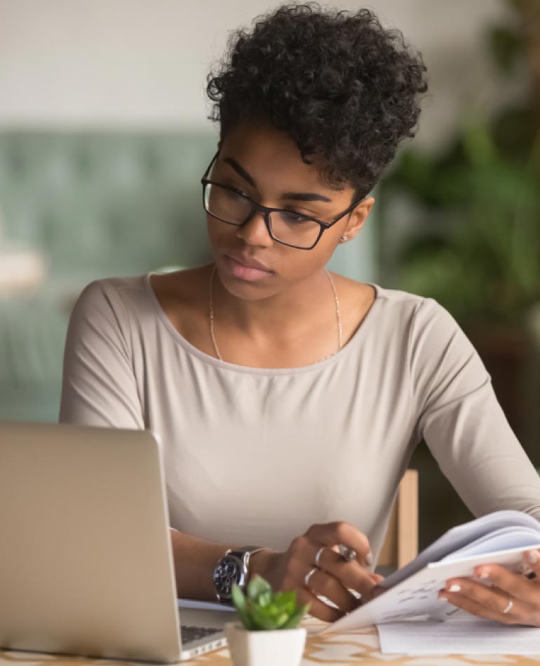 Fotografia: mulher negra sentada observa tela de notebook enquanto segura um caderno com a mão direita e tem uma caneta entre os dedos da mão esquerda. Ela possui cabelos escuros curtos e usa óculos de armação escura. Está vestida com uma blusa bege e possui um relógio no pulso direito e anéis nos dedos anelar e médio