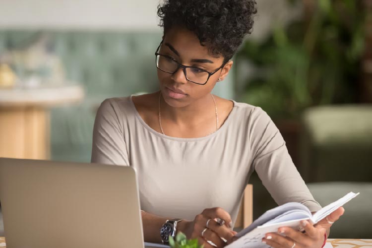 Fotografia: mulher negra sentada observa tela de notebook enquanto segura um caderno com a mão direita e tem uma caneta entre os dedos da mão esquerda. Ela possui cabelos escuros curtos e usa óculos de armação escura. Está vestida com uma blusa bege e possui um relógio no pulso direito e anéis nos dedos anelar e médio