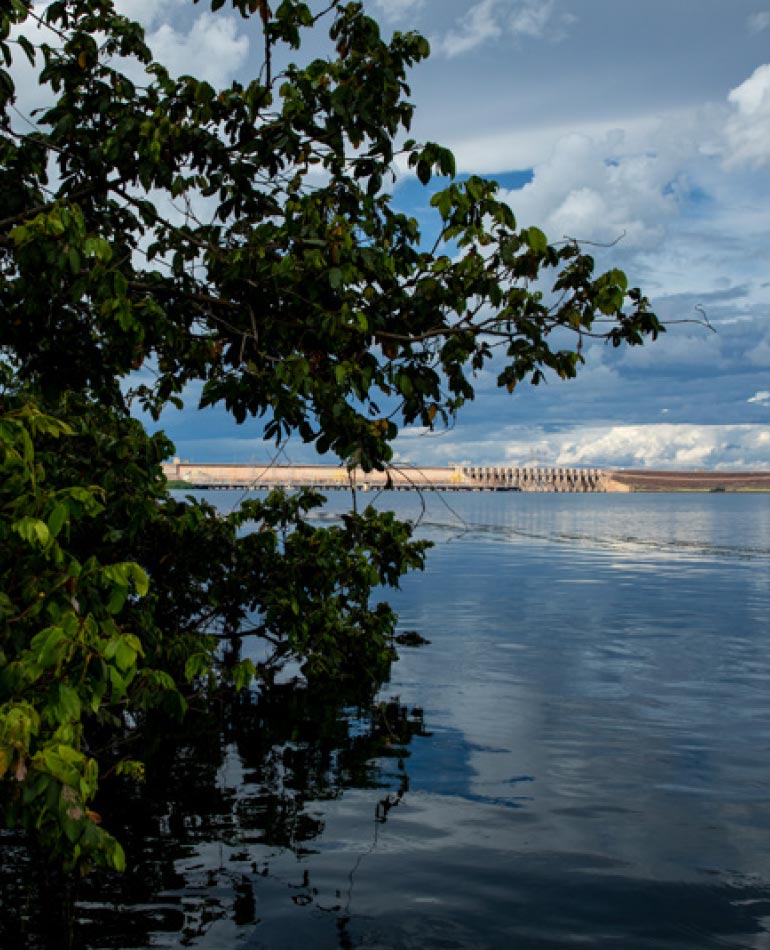 Fotografia: folhas verdes de uma árvore estão em primeiro plano, ocupando o lado esquerdo da foto. Um lago de cor azul e o céu com nuvens preechem o centro da imagem. Ao fundo, uma barragem de concreto