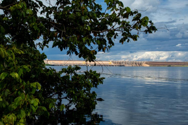 Fotografia: folhas verdes de uma árvore estão em primeiro plano, ocupando o lado esquerdo da foto. Um lago de cor azul e o céu com nuvens preechem o centro da imagem. Ao fundo, uma barragem de concreto