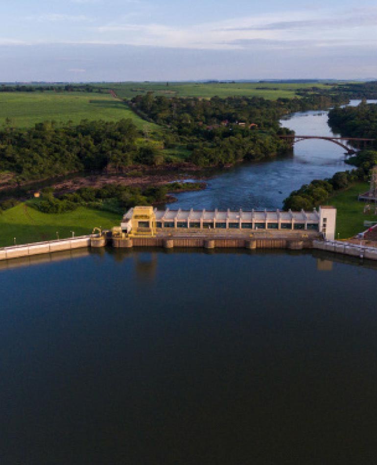 Fotografia: Na parte inferior da foto, vê-se um lago azul e a barragem de concreto de uma usina hidrelétrica. No centro da imagem, após a barragem, o rio retoma seu curso natural e as margens estão ocupadas por árvores de grande porte e áreas verdes. Ao fundo o céu azul