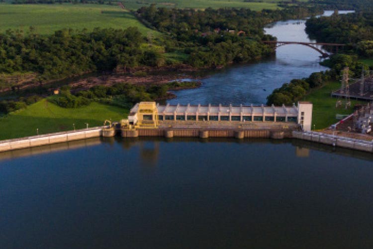 Fotografia: Na parte inferior da foto, vê-se um lago azul e a barragem de concreto de uma usina hidrelétrica. No centro da imagem, após a barragem, o rio retoma seu curso natural e as margens estão ocupadas por árvores de grande porte e áreas verdes. Ao fundo o céu azul